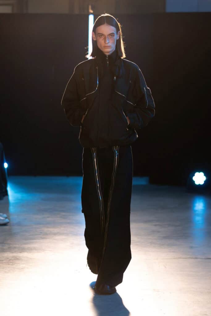 A model with straight dark hair walks a runway in a FFFPOSTAL dark, zippered jacket and wide-leg pants. The background is dimly lit with blue lights, creating a dramatic atmosphere. The model has hands in pockets.