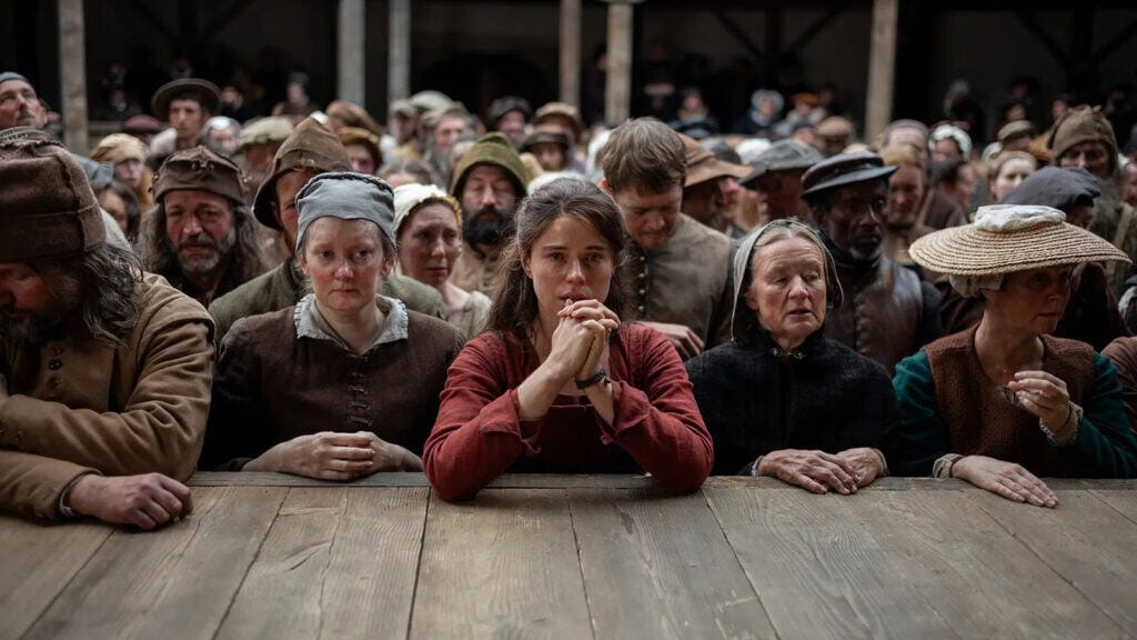 A large group of people in historical clothing stand closely together behind a wooden table, looking somber and expectant. At the center sits a young woman in red—reminiscent of Hamnet—her hands clasped, surrounded by others in muted earth tones.