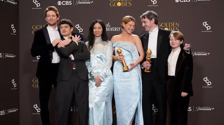 Six people dressed formally stand together smiling at the Golden Globe Awards; two women in light blue holding Hamnet trophies, and four men in suits on either side, posing in front of a branded backdrop.