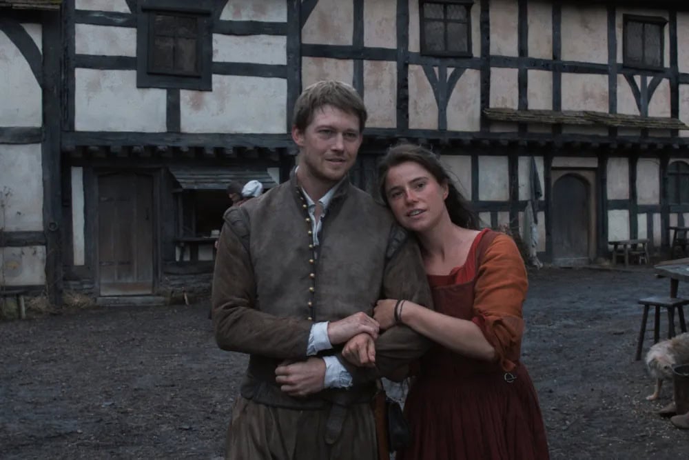 A man and woman in old-fashioned clothing stand close together outside a timber-framed building, evoking a scene from Hamnet. The woman holds the man's arm as both look toward the camera with neutral expressions in this rustic, historical setting.