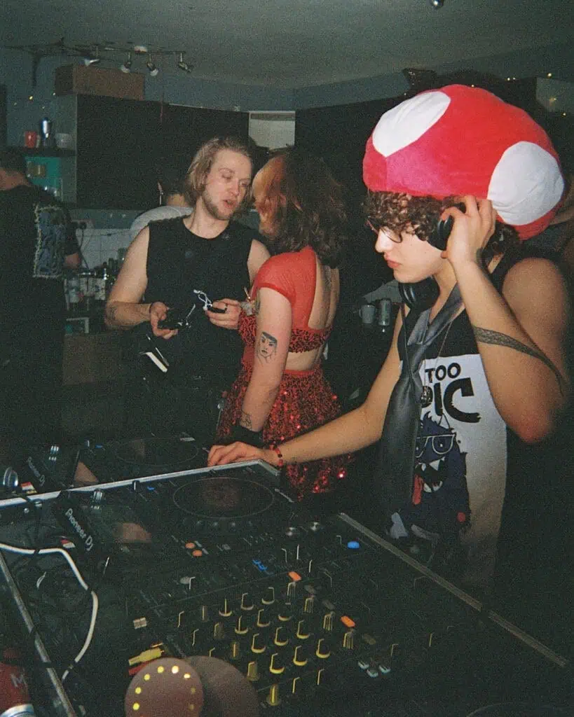 A DJ wearing a red and white mushroom hat and sunglasses mixes music at a Harringay Warehouses party. Behind the DJ, two people in black and red outfits converse in a dimly lit room filled with equipment and lights.