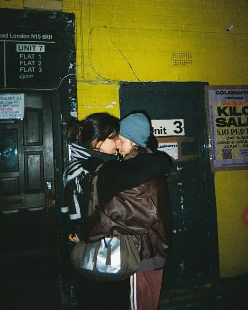 Two people embrace closely and touch noses outside a building at night in the Harringay Warehouses. They wear winter clothing and stand near a doorway lined with signs and posters on yellow and black brick walls.