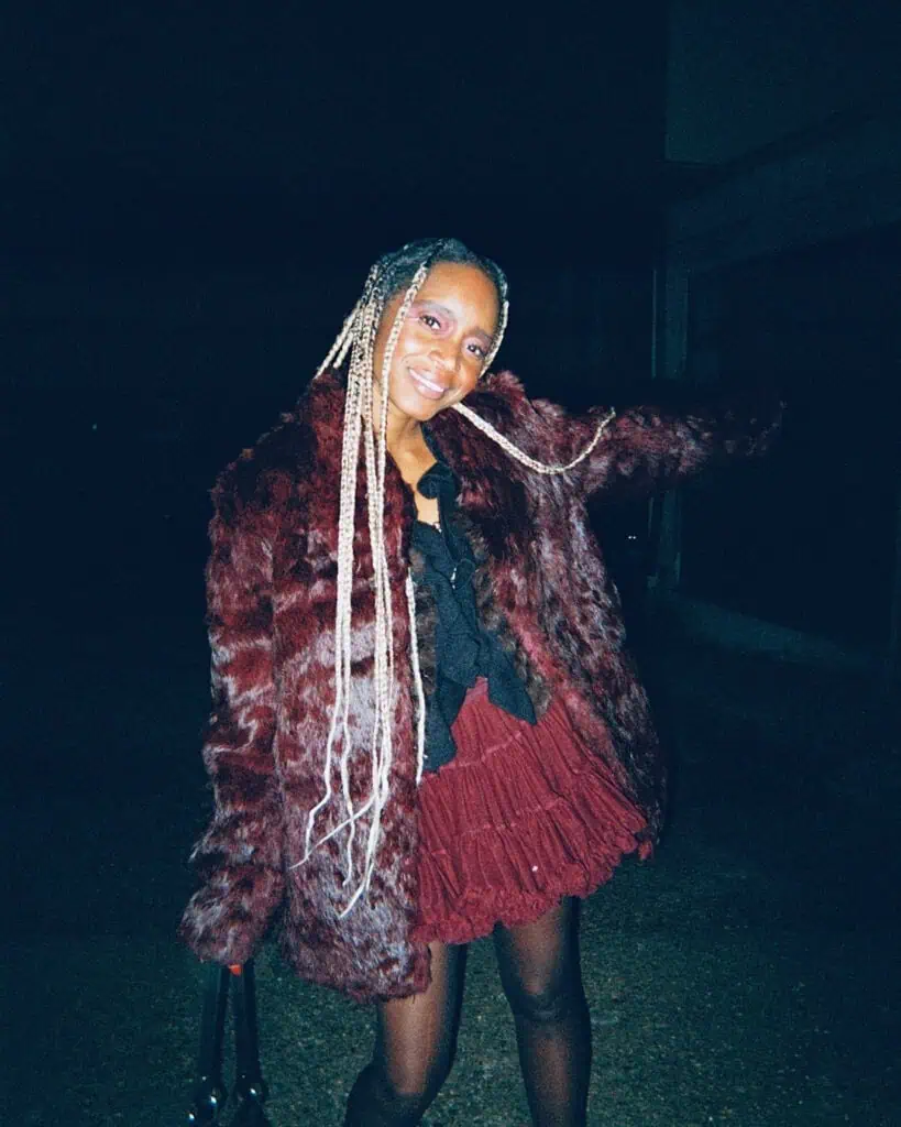 A smiling person with long, blonde braids wears a burgundy faux fur coat, a matching ruffled skirt, and dark tights. They stand outside the Harringay Warehouses at night, one arm raised while holding a bag.