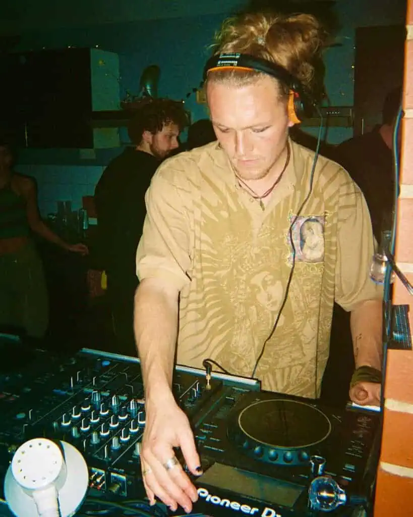 A DJ with long hair in a bun, wearing headphones and a tan graphic print shirt, adjusts controls on a Pioneer DJ mixer at a dimly lit Harringay Warehouses party. People and string lights glow in the background.