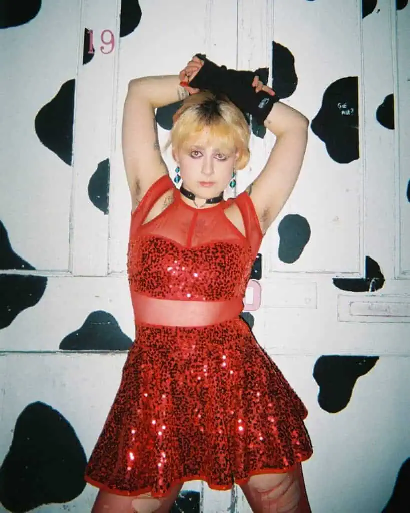 A person with short blond hair poses with arms raised, wearing a red sequined dress and black fingerless gloves, standing in front of a white door painted with black cow spots at the Harringay Warehouses.