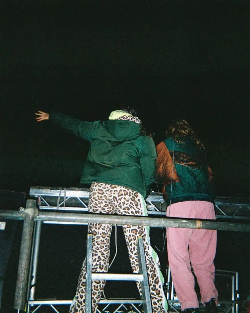 Two people stand on a metal platform at night at the Harringay Warehouses. One wears a green jacket and leopard print pants, raising an arm, while the other sports a dark jacket and pink pants. Both face away from the camera toward the dark sky.