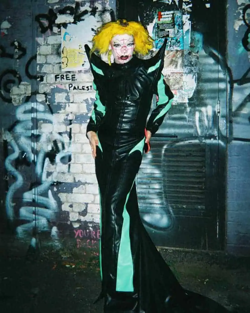 A person in a dramatic black and green gown with exaggerated shoulders and vibrant yellow hair poses against a graffiti-covered wall at the Harringay Warehouses. Their face is painted with bold, artistic makeup. A Free Palestine message is visible on the wall.