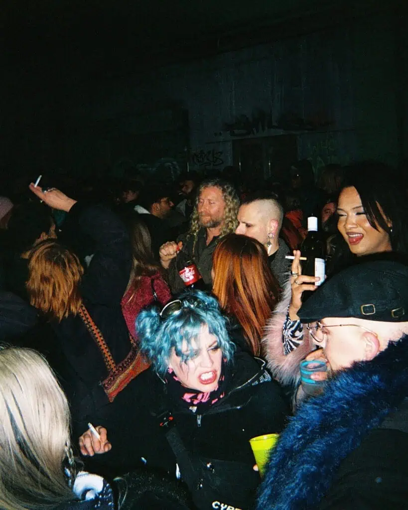 A lively crowd at a night party in the Harringay Warehouses, many holding drinks and cigarettes. One person with bright blue hair and dramatic makeup stands out in the foreground as others laugh and interact in the dimly lit scene.