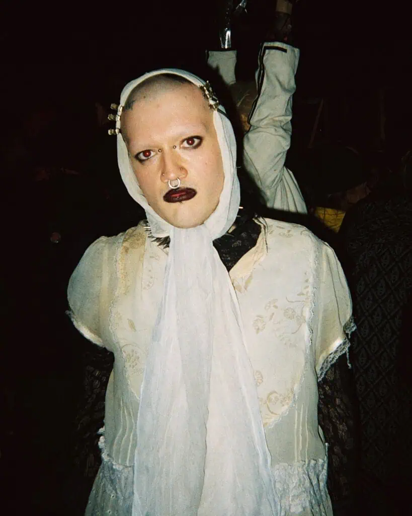 A person with a shaved head, bold dark lipstick, dramatic eye makeup, and facial piercings wears a cream-colored vintage dress and headscarf, standing in the Harringay Warehouses crowd at night, staring intensely at the camera.