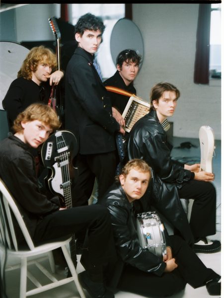 Six young men dressed in black pose with musical instruments, including a guitar, drum, keyboard, bass guitar, and tambourine, in a brightly lit indoor space with white walls and red curtains, ready to perform songs from their latest albums.
