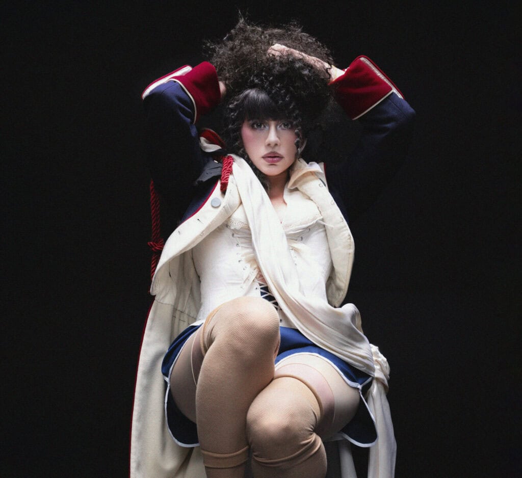 KÁRYYN in theatrical makeup and costume, wearing a dark jacket with red cuffs, a white corset, blue skirt, and beige stockings, sits with hands in curly hair against a dark background.