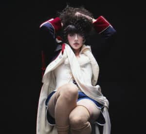 KÁRYYN in theatrical makeup and costume, wearing a dark jacket with red cuffs, a white corset, blue skirt, and beige stockings, sits with hands in curly hair against a dark background.