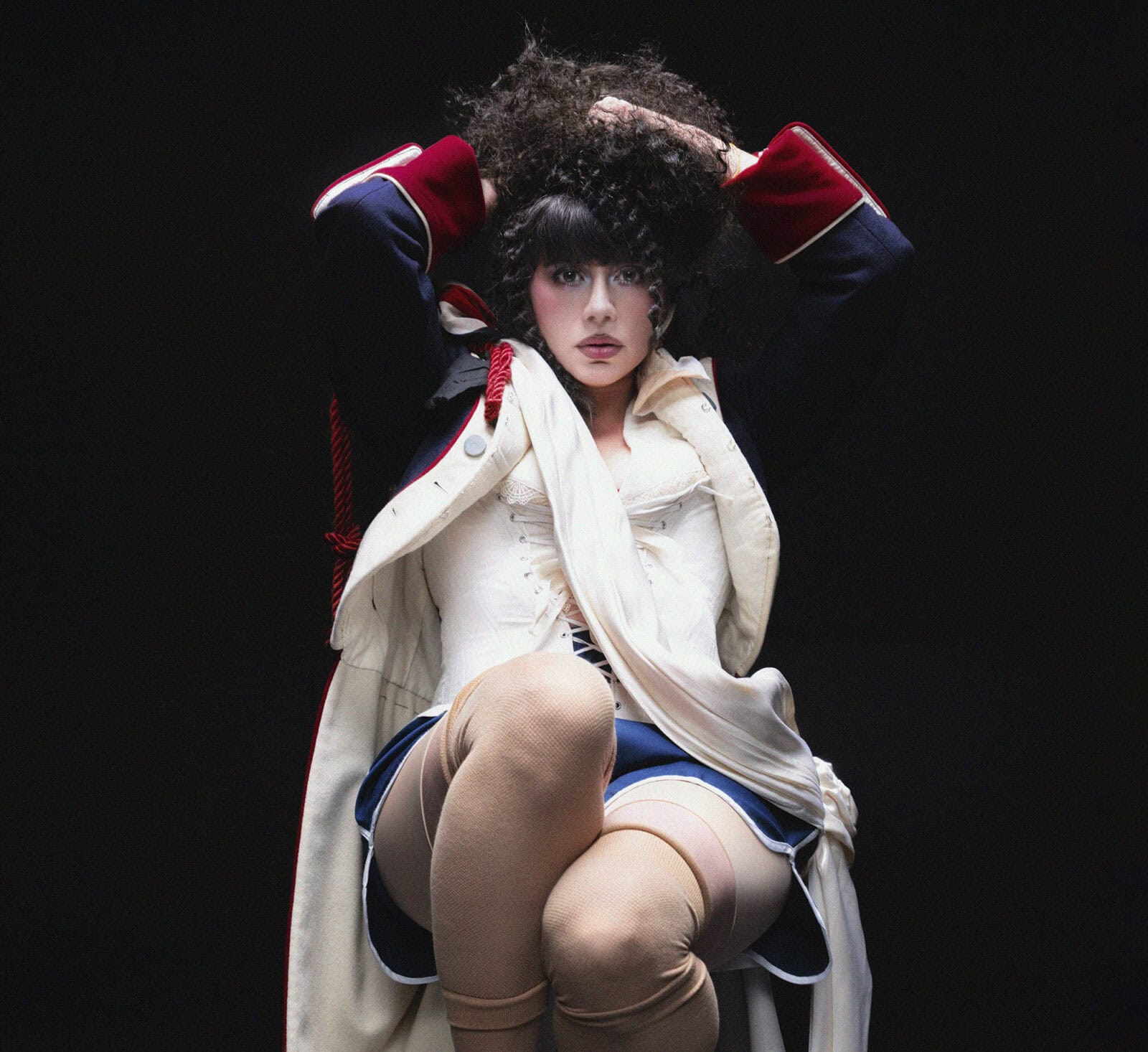 KÁRYYN in theatrical makeup and costume, wearing a dark jacket with red cuffs, a white corset, blue skirt, and beige stockings, sits with hands in curly hair against a dark background.