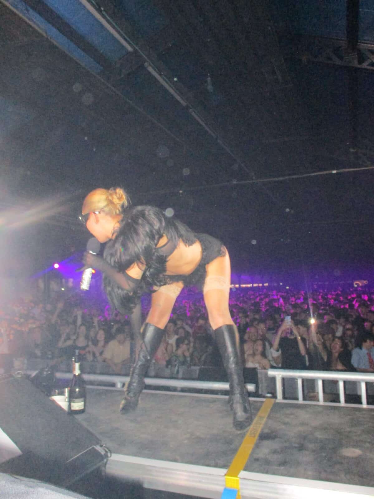 A performer in black boots, shorts, and a furry top bends forward on stage at 2much Festival, holding a microphone and facing a large crowd under colorful stage lights. A champagne bottle rests near the stage’s edge.