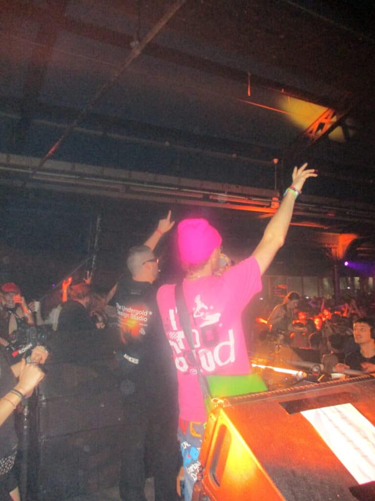 A performer in a pink shirt and beanie faces a cheering crowd on stage at 2much Festival, raising one arm. Another person stands nearby, also gesturing to the audience as colorful stage lights brighten the dimly lit venue.