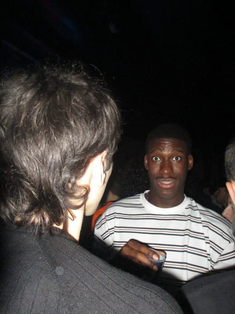 Two people are talking in a dimly lit nightclub at the 2much Festival. One person, facing the camera and holding a drink, looks surprised, while the other is seen from behind with only the back of their head visible.