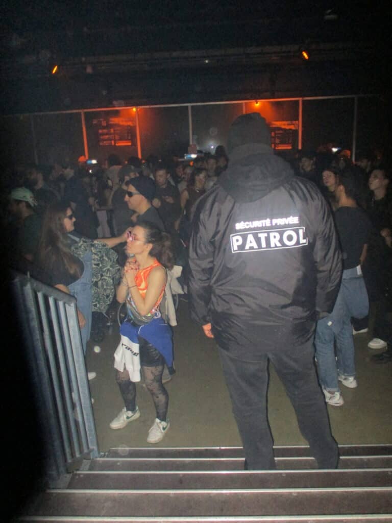A security guard in a black jacket labeled Sécurité Privée Patrol stands near a lively crowd dancing and socializing at the 2much Festival in a dimly lit club or concert venue.