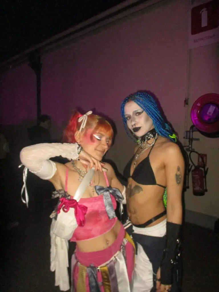 Two people pose at the 2much Festival, wearing colorful, edgy outfits with bold accessories. One has orange-pink hair in pigtails; the other rocks blue braids and striking makeup. A fire extinguisher is visible in the background.
