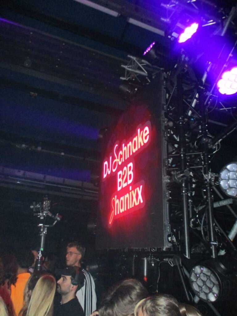 A large illuminated sign reads DJ Schnake B2B Shanixx in red letters at a crowded indoor 2much Festival event, with people standing nearby and stage lights overhead.