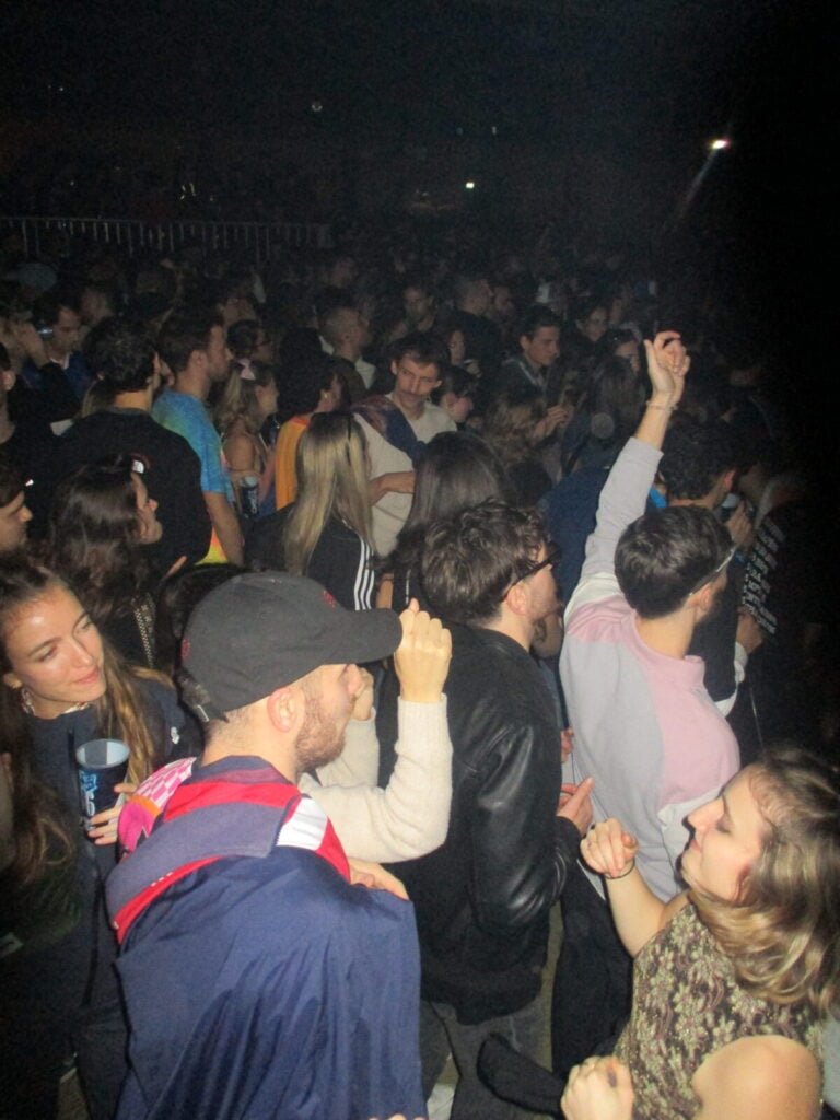 A large crowd of young people dance and socialize at the lively 2much Festival, with some raising their hands in the air and others talking amongst themselves. The atmosphere appears energetic and crowded.