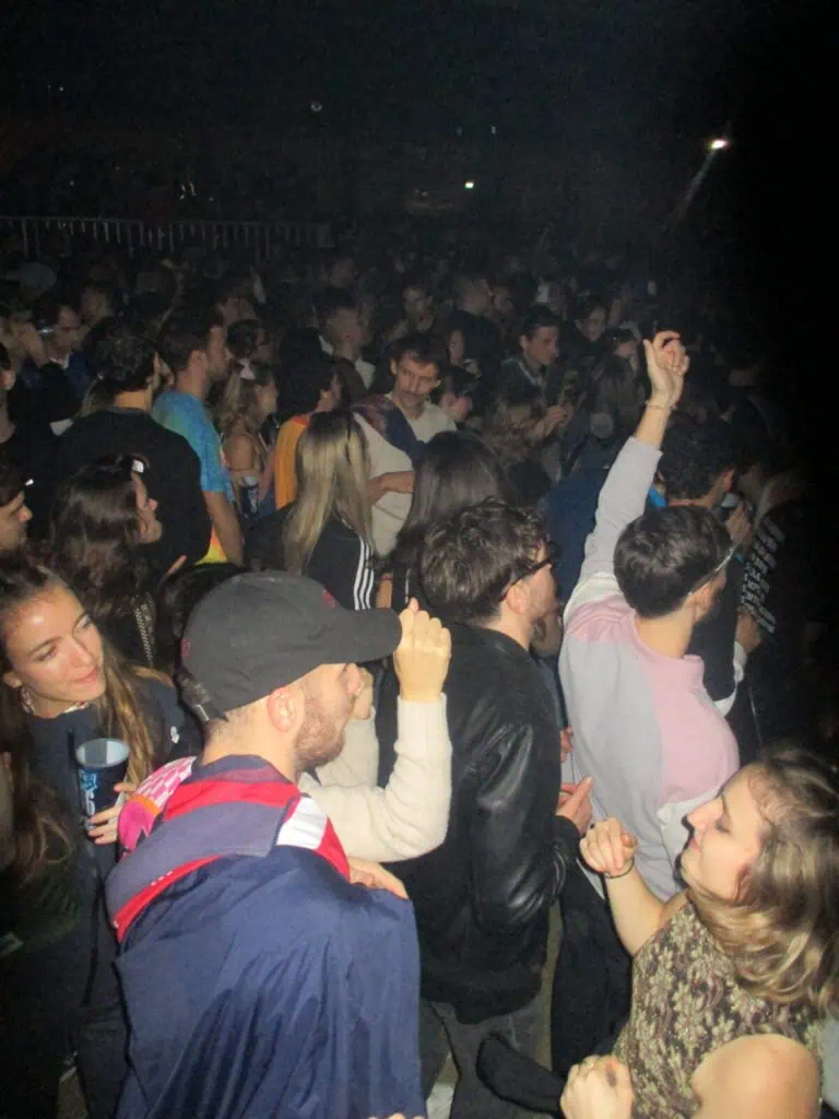 A large crowd of young people dance and socialize at the lively 2much Festival, with some raising their hands in the air and others talking amongst themselves. The atmosphere appears energetic and crowded.