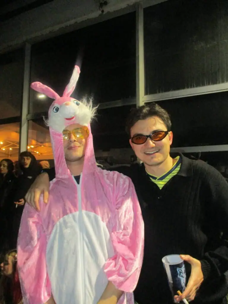 Two people posing together at 2much Festival—one in a pink bunny costume with a rabbit head hood and sunglasses, the other in dark clothing with orange sunglasses, smiling and holding a cup. People and lights fill the background.