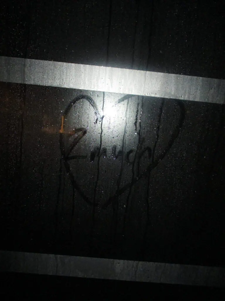 A foggy window with a heart shape and the word Amor drawn on it captures the spirit of 2much Festival. Two horizontal white stripes cross the glass, while a bright light from behind highlights the heartfelt drawing.