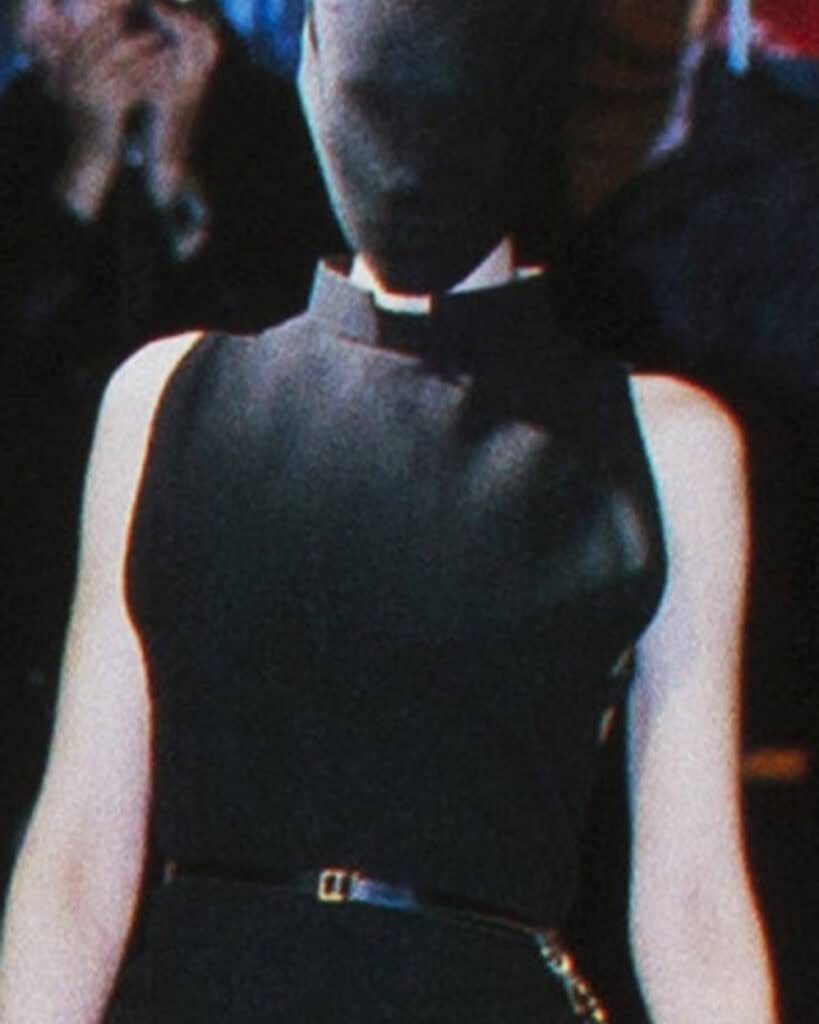 A person wearing a sleeveless black dress and a matching black head covering stands facing away from the camera. The background is blurred, showing a seated audience.