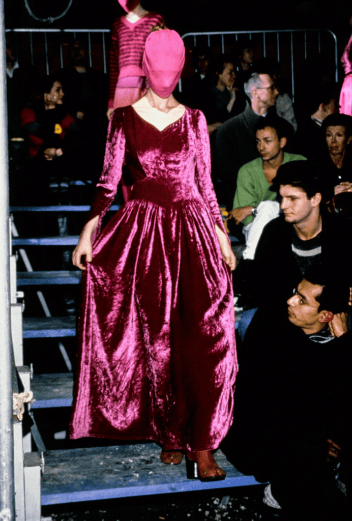 A model wearing a vibrant pink velvet gown reminiscent of Margiela AW95 and a matching pink face-covering walks down steps at a fashion show, with audience members seated closely on either side.