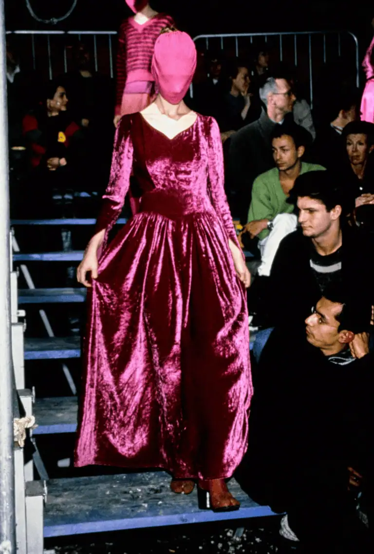A model wearing a vibrant pink velvet gown reminiscent of Margiela AW95 and a matching pink face-covering walks down steps at a fashion show, with audience members seated closely on either side.