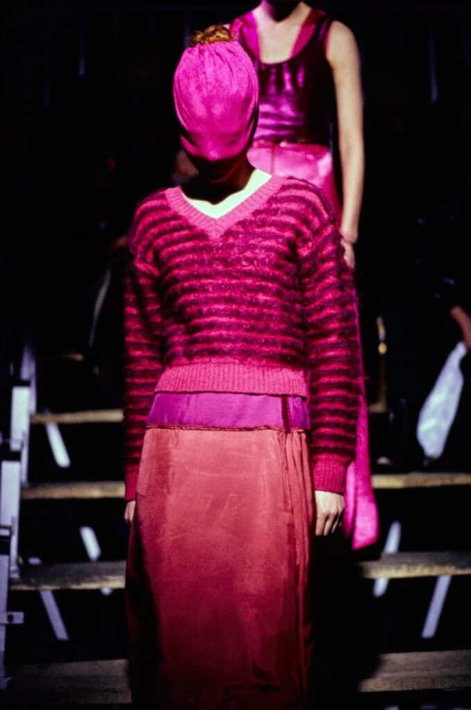 A model wearing a pink striped sweater, a long pink skirt, and a wrapped pink head covering walks a dimly lit runway, with another model in a pink dress visible in the background.