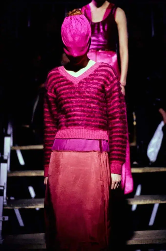 A model wearing a pink striped sweater, a long pink skirt, and a wrapped pink head covering walks a dimly lit runway, with another model in a pink dress visible in the background.