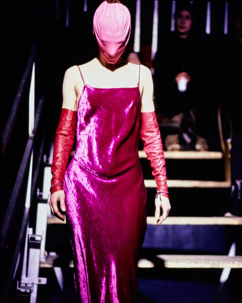 A person walks down stairs wearing a shiny fuchsia slip dress, long matching gloves, and a pink head covering that obscures their face. The background shows someone seated on the steps.