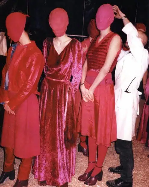 Three people dressed in various shades of red clothing stand side by side with their faces fully covered by red fabric, while a person in a white coat adjusts one of their coverings.