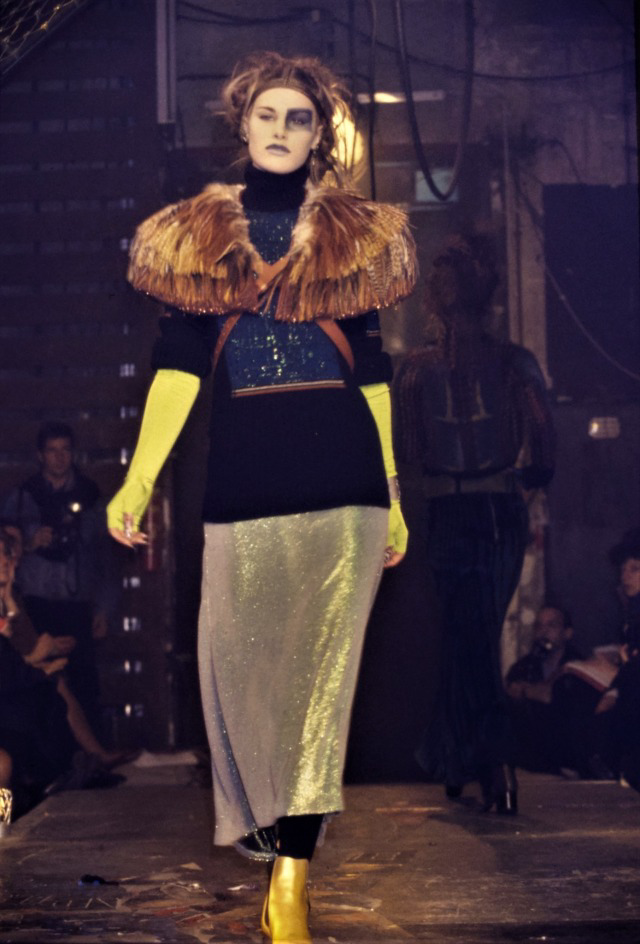 A model walks a dimly lit runway wearing a dramatic outfit with a fur-like shoulder piece, black top, long yellow gloves, sparkly silver skirt, and yellow boots. The setting appears industrial, with a seated audience in the background.