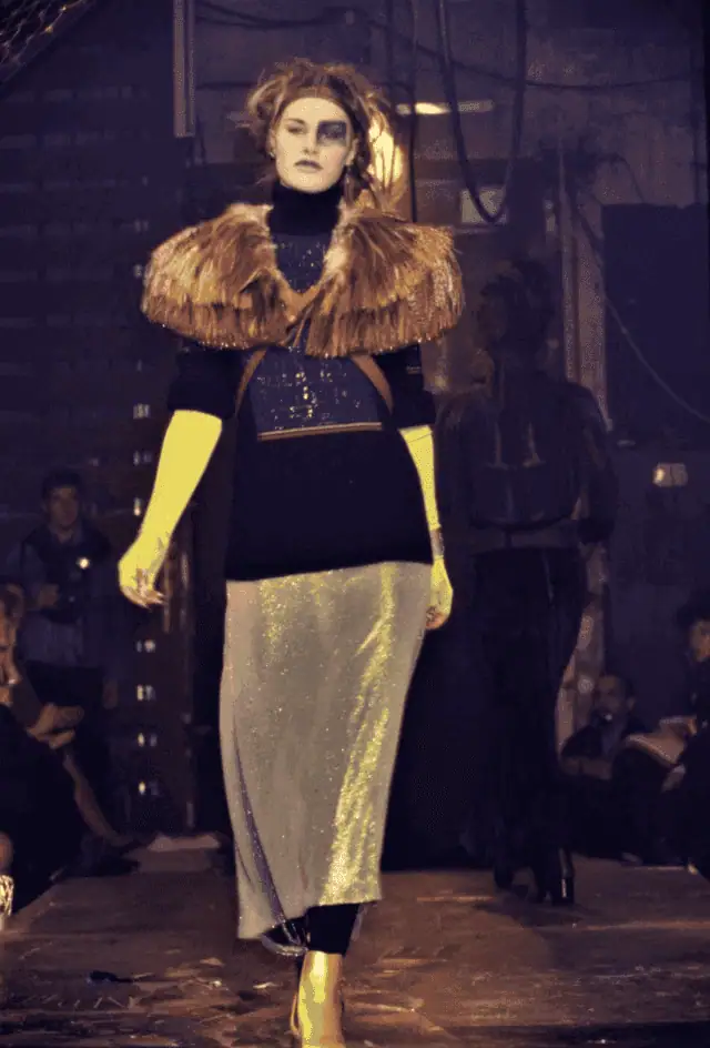 A model walks a dimly lit runway wearing a dramatic outfit with a fur-like shoulder piece, black top, long yellow gloves, sparkly silver skirt, and yellow boots. The setting appears industrial, with a seated audience in the background.