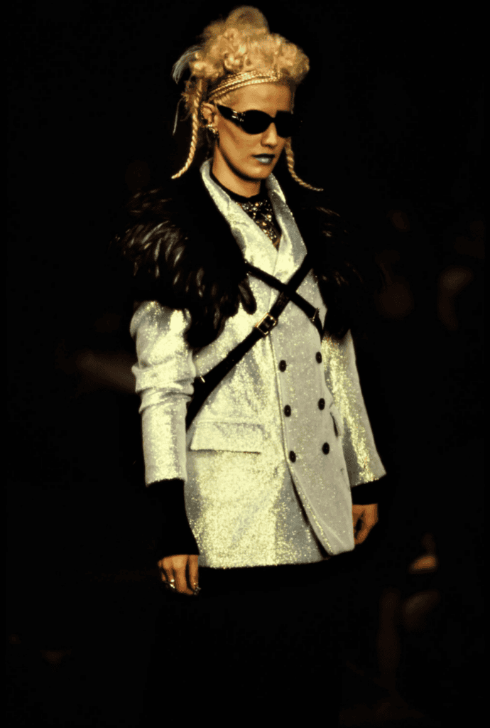 A model walks the runway wearing a shiny silver double-breasted blazer with black fur shoulders, black harness, dark sunglasses, light lipstick, and styled blonde hair adorned with braids and accessories.