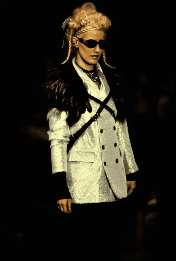 A model walks the runway wearing a shiny silver double-breasted blazer with black fur shoulders, black harness, dark sunglasses, light lipstick, and styled blonde hair adorned with braids and accessories.