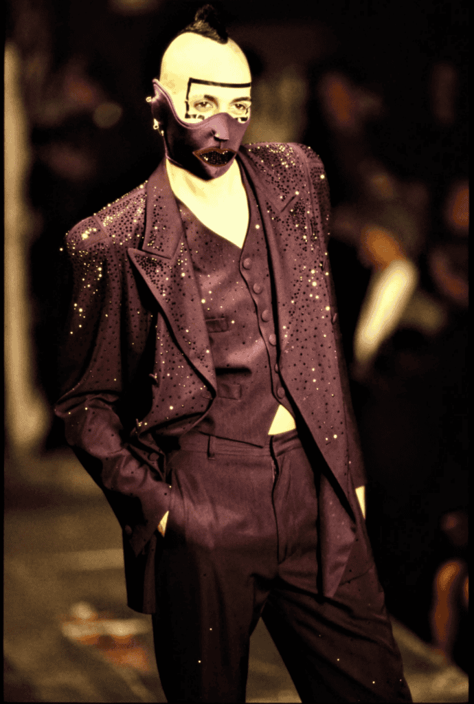 A model wearing a purple, rhinestone-studded suit with a matching vest walks the runway. Their face is painted pale with bold geometric makeup, and they wear a dramatic purple mask resembling an open mouth.