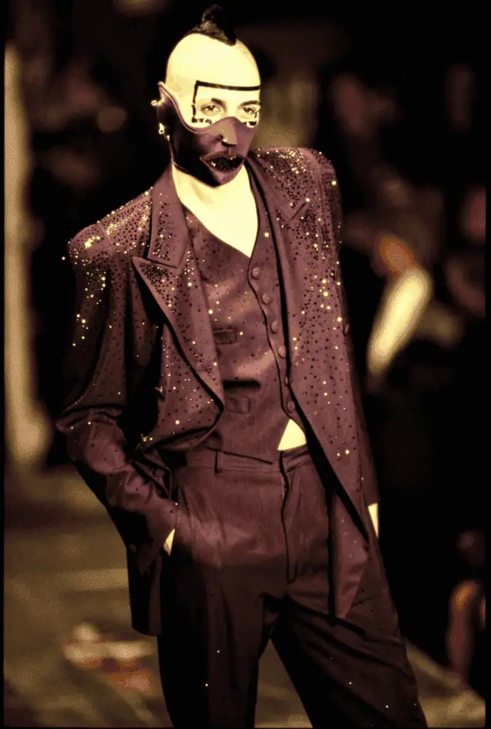 A model wearing a purple, rhinestone-studded suit with a matching vest walks the runway. Their face is painted pale with bold geometric makeup, and they wear a dramatic purple mask resembling an open mouth.