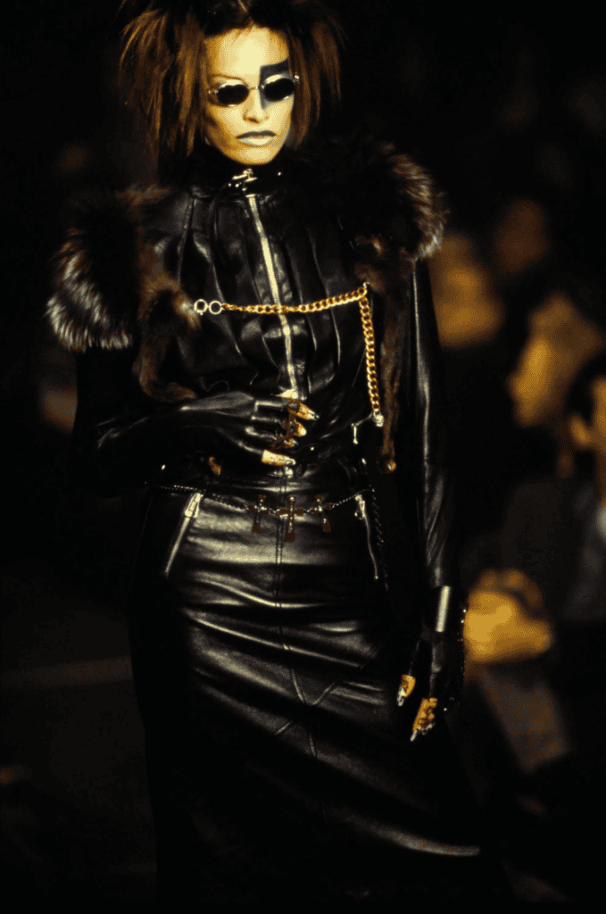 A model wearing sunglasses, a black leather outfit with fur accents, gloves, and gold chain details walks the runway against a dark, blurred background.