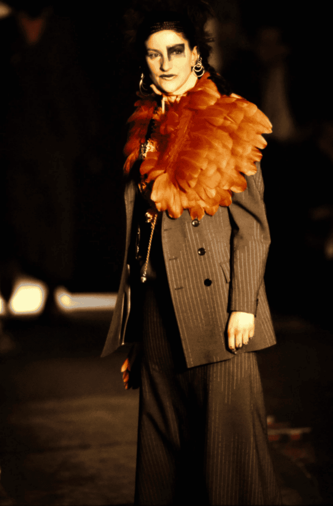 A model walks a dimly lit runway wearing a gray pinstripe suit and wide-legged trousers, accessorized with a dramatic orange feathered collar and bold, dark eye makeup.