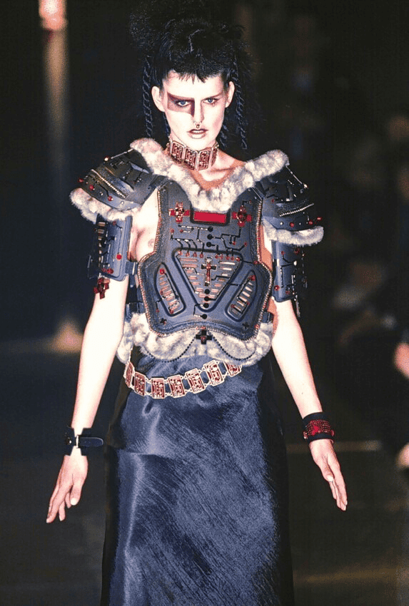 A model walks the runway wearing futuristic, armored shoulder pads with fur trim, a structured chest plate, bold makeup, and a long black skirt, blending sci-fi and tribal elements in a dramatic fashion look.