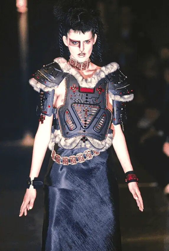 A model walks the runway wearing futuristic, armored shoulder pads with fur trim, a structured chest plate, bold makeup, and a long black skirt, blending sci-fi and tribal elements in a dramatic fashion look.