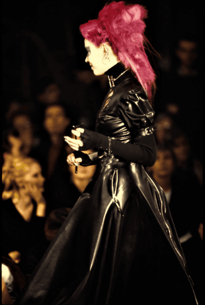 A person with bright pink hair styled in a tall, voluminous mohawk wears a dramatic, shiny black leather dress with long sleeves, walking past a blurred audience at a fashion event.
