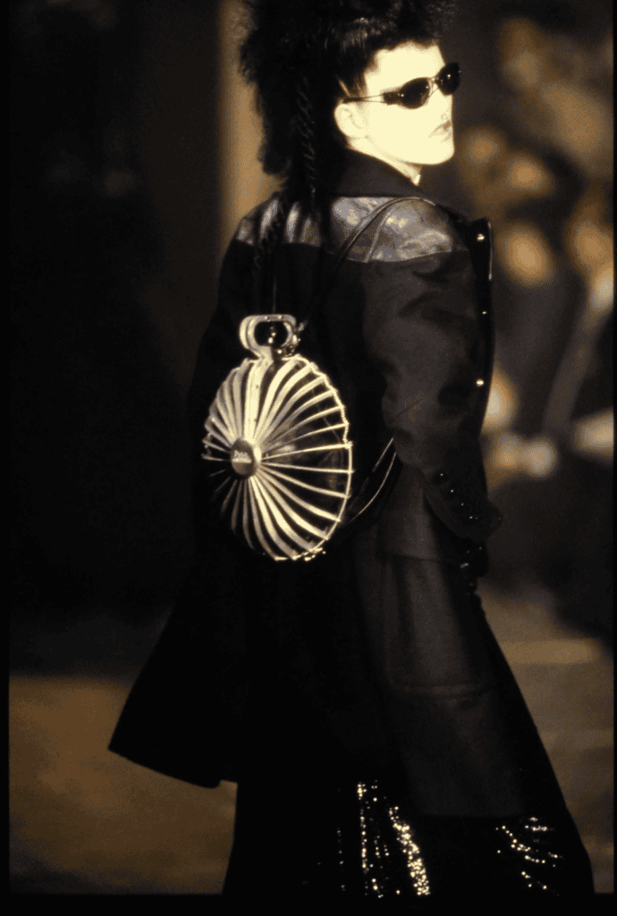 A model wearing a black coat, dark sunglasses, and a spiked hairstyle walks a runway carrying a unique, circular silver bag with a radiating design on their back.
