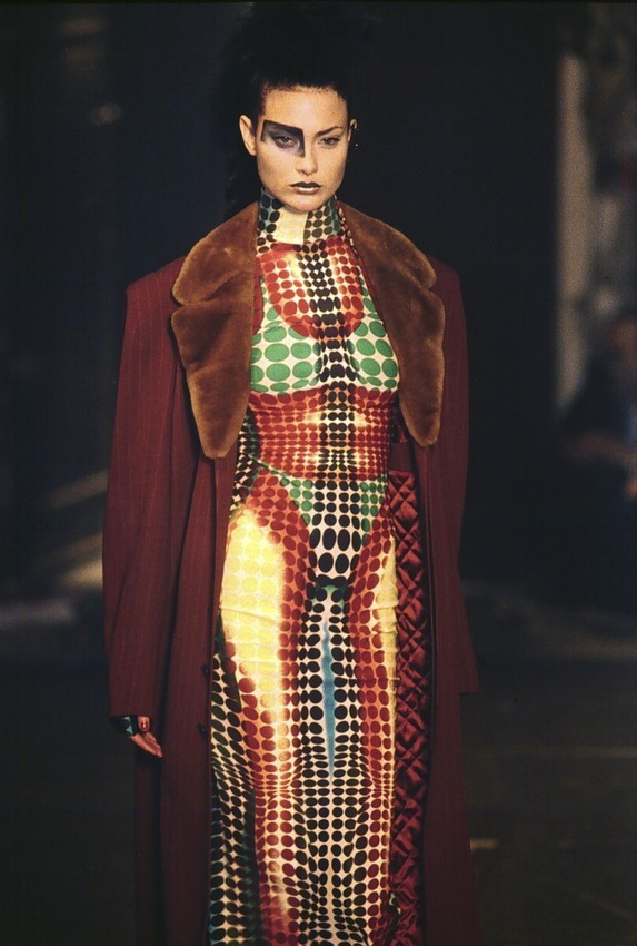 A model walks the runway wearing a bold, colorful dress with geometric dot patterns in red, green, black, and yellow, paired with a deep red coat with a large brown fur collar. She has dramatic makeup and an intense expression.