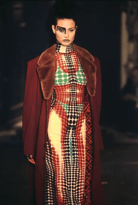 A model walks the runway wearing a bold, colorful dress with geometric dot patterns in red, green, black, and yellow, paired with a deep red coat with a large brown fur collar. She has dramatic makeup and an intense expression.
