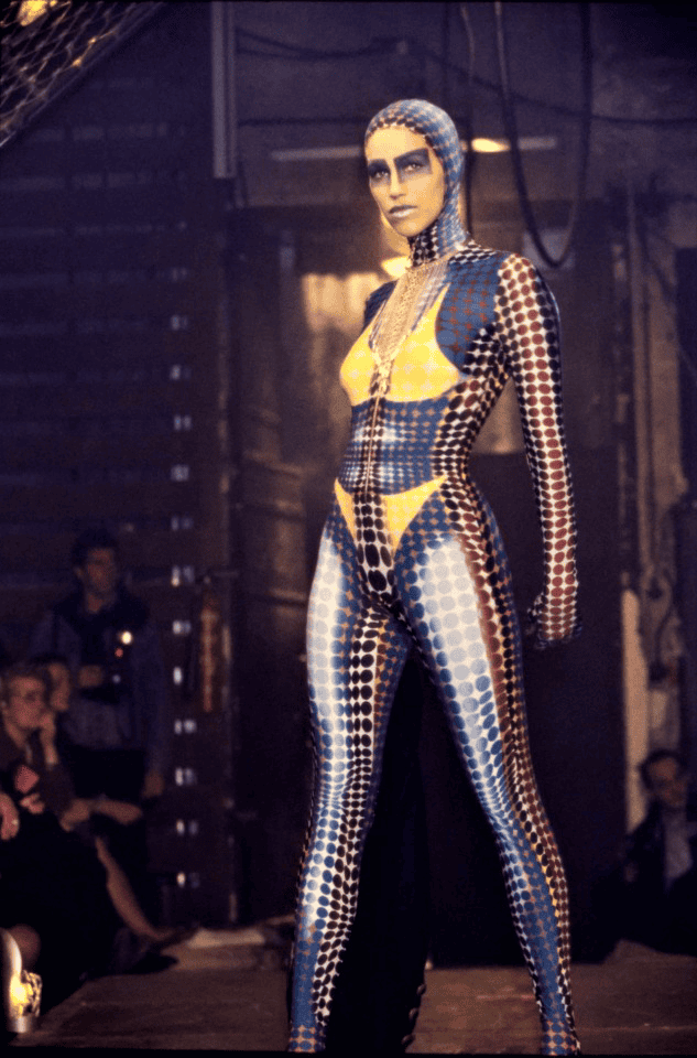 A model walks a runway wearing a colorful, form-fitting bodysuit with a geometric, dot pattern in blue, yellow, red, and white. The suit covers her head, and she stands confidently in an industrial setting.