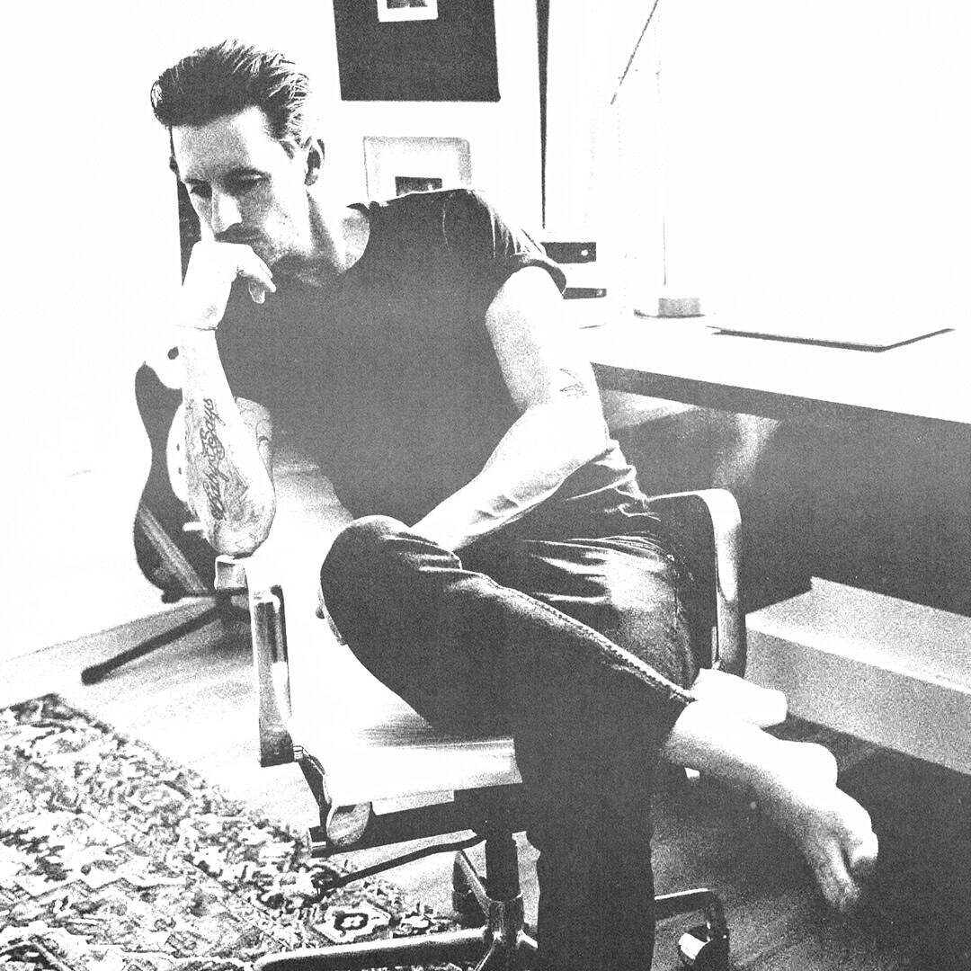 A person with tattoos sits barefoot on a chair in a modern, bright room, leaning forward with their hand on their chin, appearing thoughtful—perhaps reflecting on ideas for their cold team. A desk, framed art, and patterned rug are visible in the background.